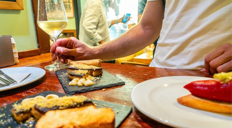 A man at a wooden bar counter with a glass of txakoli and seared foie pintxos on slate