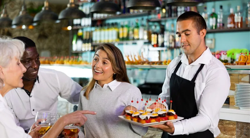 A waiter carrying a tray of pintxos to guests standing at a busy Basque bar