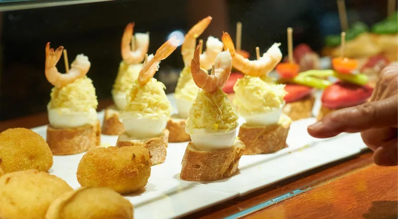 A hand reaching towards prawn and egg pintxos in a lit glass counter case